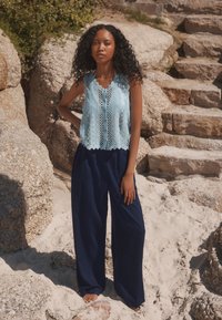 Light blue lace sleeveless top with scalloped hem, paired with loose navy trousers. Setting: sandy beach, stone background.