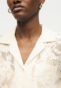 Κοντινό πλάνο ενός ατόμου που φοράει μια κρεμ δαντελένια μπλούζα με γιακά και λουλουδάτα σχέδια, καθώς και χρυσούς κρίκους σκουλαρίκια.