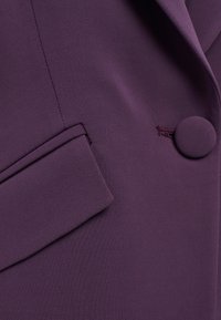 Tessuto in velluto viola intenso con una superficie liscia, presenta un unico bottone rotondo e un piccolo dettaglio della tasca inclinata.