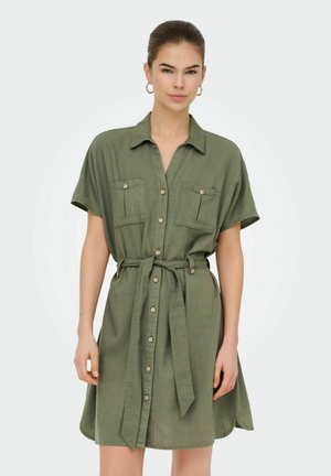 Kvinde iført en kortærmet olivengrøn kjole med knapper, brystlommer og bælte bundet i taljen, stående foran en ensfarvet baggrund.