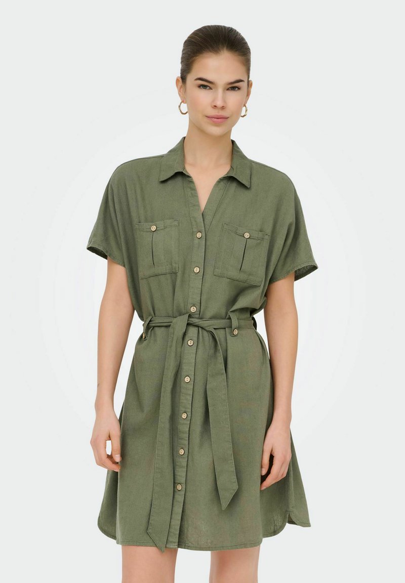 Femme portant une robe boutonnée manches courtes de couleur olive avec des poches poitrine et une ceinture nouée à la taille, debout devant un fond uni.