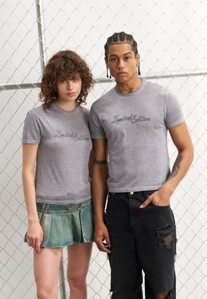 T-shirts gris à manches courtes avec le texte "Édition Limitée". Une personne porte une jupe en denim plissée ; l'autre porte un jean noir déchiré.