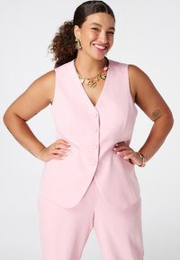 Lichtroze mouwloos vest met een V-hals en vijf knopen, gecombineerd met bijpassende broek. Het model is accessorized met bloemenoorbellen en sieraden.