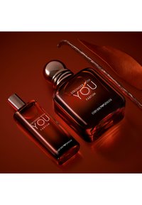 Due bottiglie di profumo ambra scuro etichettate "Stronger With You Parfum" di Emporio Armani accanto a una bacca di vaniglia e una foglia arancione essiccata.