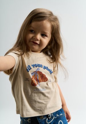 Tricou bej din bumbac cu un grafic cu un croissant și text albastru. Asortat cu blugi din denim care au un model alb scris de mână.