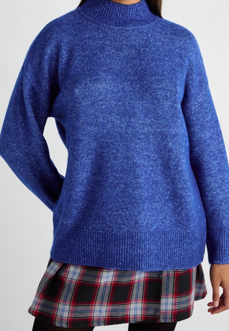 Femme portant un pull en tricot bleu à col montant et une jupe à carreaux rouge, grise et noire, sur un fond uni.