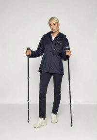 Veste imperméable marine avec capuche, poches zippées et poignets élastiques, associée à un pantalon sombre et des baskets blanches, tenant des bâtons de trekking.