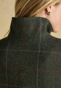 Collo in lana a scacchi verde e grigio con collo alto, caratterizzato da sottili linee a rete rosse e viola. Orecchino a cerchio dorato visibile.