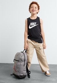 Pilkas kuprinė su juodais akcentais, kuriame yra raudonas Jordan logotipas. Vaikas dėvi juodą Nike marškinėlį be rankovių ir smėlio spalvos krovinius, šypsosi.