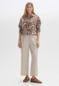 Brown and beige patterned button-up shirt, paired with light gray loose trousers and brown sandals. Smooth fabric, cropped design.