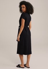 Abito midi nero con maniche corte, vita aderente con cintura e tessuto morbido. Modella che indossa sandali neri piatti, vista da dietro.