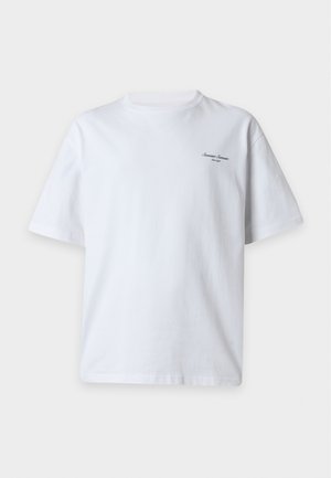 Homme debout portant un t-shirt blanc ample à manches courtes, un short noir large jusqu'aux genoux et des bottines noires sur un fond blanc uni.