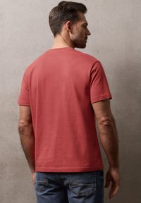 Homme vu de dos portant un t-shirt rouge uni à manches courtes et un jean bleu, devant un mur gris clair texturé.