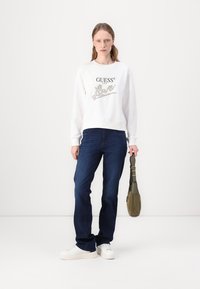 Felpa bianca con il logo “GUESS” e il testo scintillante “Love”, abbinata a jeans blu scuro e sneakers bianche. Borsa verde oliva tenuta con una mano.