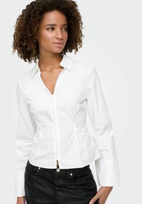 Chemise blanche à col en V, design ajusté, manches longues et texture lisse, portée avec un jean noir.