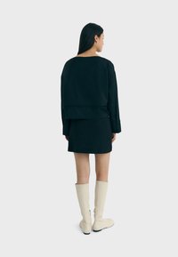 Schwarzer, verkürzter Pullover, der über einem schwarzen Rock getragen wird, kombiniert mit kniehohen, cremefarbenen Stiefeln. Glattes Material mit minimalistischer Design.