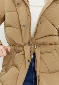 Puffer jacket in beige with a quilted texture, a zipper closure, snap buttons, and an adjustable drawstring at the waist.