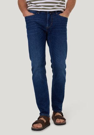 Blue denim jeans with a slim fit, featuring front pockets, belt loops, and subtle stitching details. Worn with brown sandals.