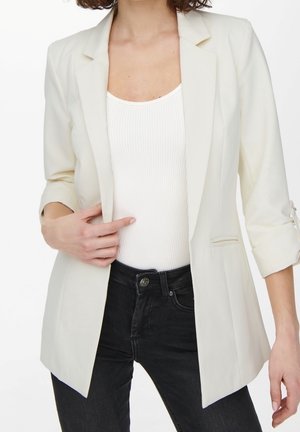 Femme portant un blazer crème aux manches retroussées sur un débardeur blanc côtelé et un jean taille haute noir, la main touchant le devant du blazer.