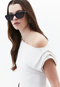 Occhiali da sole neri oversize con montatura quadrata, indossati con una maglietta bianca a spalle scoperte con maniche drappeggiate. Lunghi capelli castani cadono delicatamente.