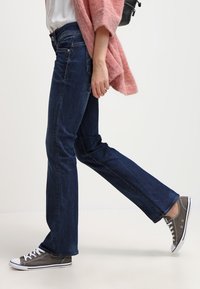 Dark blue denim jeans with a straight leg fit, paired with gray low-top sneakers and a loose pink knit cardigan.