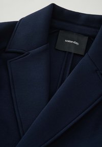 Marineblazer mit glatter Textur, Reverskragen und einer einzelnen Tasche. Das Etikett trägt die Aufschrift "irgendwann". Sichtbare Nahtdetails entlang des Kragens.