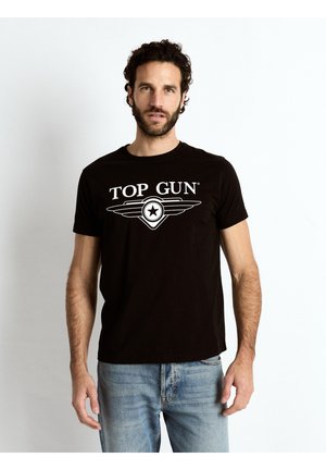 Mężczyzna z kręconymi włosami i brodą, ubrany w czarną koszulkę "Top Gun" i niebieskie dżinsy, stojący na tle jednolitego jasnego tła.