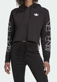 Svart kort hoodie med vitt Adidas-logotyp på bröstet och grafisk text längs ärmarna. Har en dragsko-huva och ribbade ärmslut.