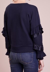 Sudadera azul marina con mangas de satén con volantes, escote redondo y corte relajado. Combinada con pantalones vaqueros azules, que muestran una textura de tela suave.