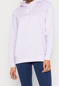 Person som bär en ljuslila Nike-hoodie med snoddar och marinblå leggings, stående mot en enkel vit bakgrund.