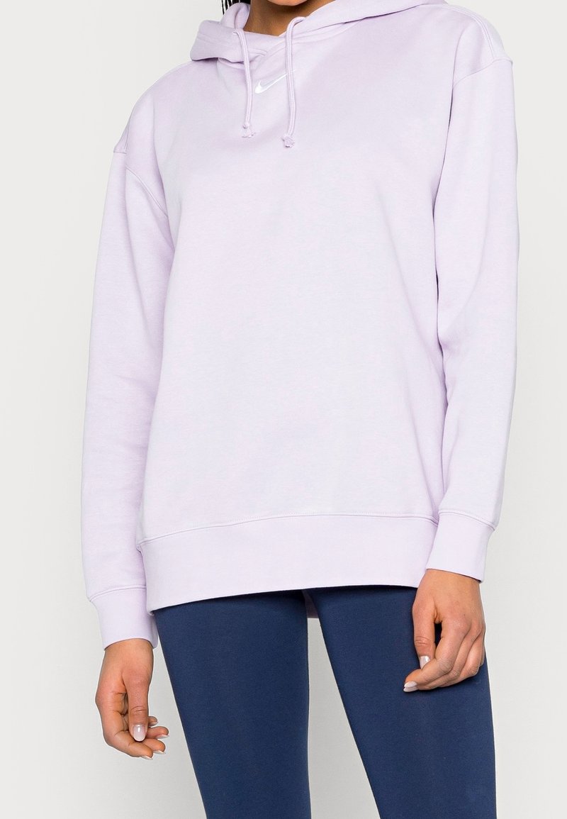 Person som bär en ljuslila Nike-hoodie med snoddar och marinblå leggings, stående mot en enkel vit bakgrund.