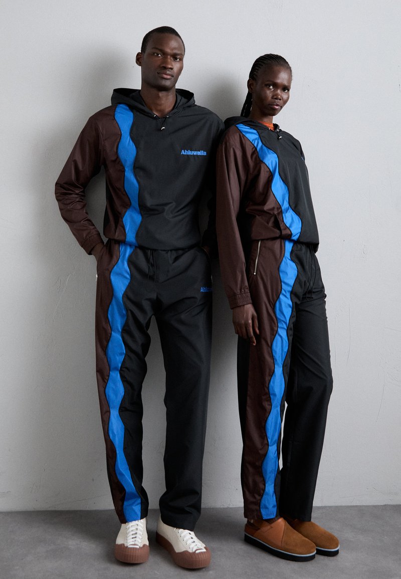 Deux mannequins portent des survêtements de sport coordonnés en tissu noir et marron avec des rayures d'accent bleu. Leur chaussure contraste avec des tons clairs et foncés.