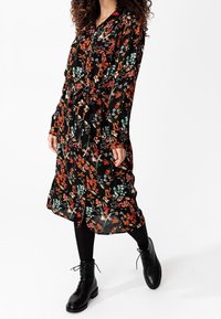 Abito a stampa floreale con sfondo nero, caratterizzato da colori vivaci come rosso e verde, maniche lunghe, una vita legata e una silhouette fluida.