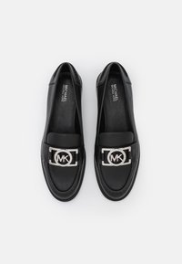 Mocassins en cuir noir avec un détail en métal argenté représentant le logo MK sur le devant. Texture lisse, bout arrondi et design bas.