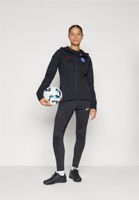 Svart Nike hettegenser med glidelås og logo, kombinert med svarte teksturerte bukser og svarte sportsko. Spilleren holder en hvit og blå fotball.
