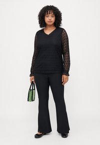 Μαύρη δαντελένια μπλούζα με μακριά μανίκια, V-αλυσιδωτή λαιμόκοψη και λουλουδάτα σχέδια. Συνδυασμένη με μαύρο φαρδύ παντελόνι. Αξεσουάρ με πράσινη τσάντα με χάντρες.