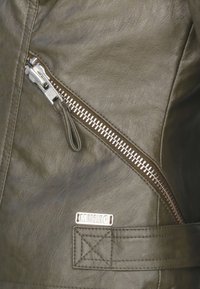 Olivgrüne Lederjacke mit einem diagonal verlaufenden silbernen Reißverschluss, metallischen Beschlägen und einem rechteckigen Etikett mit der Prägung "MUSTANG."