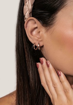 Close-up of a woman's ear with two small rose gold hoop earrings, wearing a beige braided headband and touching her face with manicured nails.