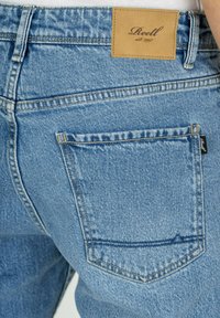 Jeans de mezclilla de color azul claro con una etiqueta de cuero visible que dice "Reell est. 1997," con un bolsillo trasero y sutiles detalles de desgaste.