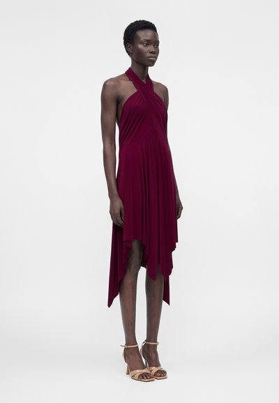 Ψηλό άτομο που φοράει αμάνικο, βαθύ κόκκινο φόρεμα χαλτέρ με ασύμμετρο τελείωμα και μπεζ πέδιλα με λουράκι στον αστράγαλο με τακούνι, στέκεται μπροστά σε ένα μονόχρωμο φόντο.