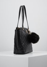 Sac cabas matelassé noir avec motif en relief, texture brillante, quincaillerie dorée et un pompon en fourrure noire détachable.