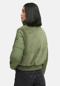 Green puffer jacket with a soft, shaggy texture on the back, ribbed hem, and smooth sleeves. Model facing away, wearing dark jeans.