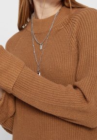 Eine Frau trägt einen gerippten braunen Pullover und zwei geschichtete silberne Halsketten mit kleinen Anhängern, sichtbar von Hals bis zur Mitte des Oberkörpers.
