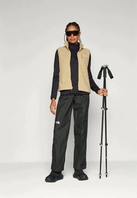 Colete de polar bege sobre uma blusa de manga longa preta, combinado com calças e sapatos pretos à prova de água. Segurando um bastão de trekking.