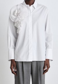 Camicia bianca con bottoni caratterizzata da un grande accento floreale bianco plissettato sulla spalla sinistra. Maniche lunghe e vestibilità comoda completano il design.