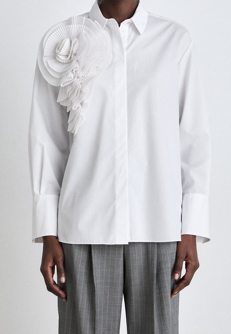 Camicia bianca con bottoni caratterizzata da un grande accento floreale bianco plissettato sulla spalla sinistra. Maniche lunghe e vestibilità comoda completano il design.