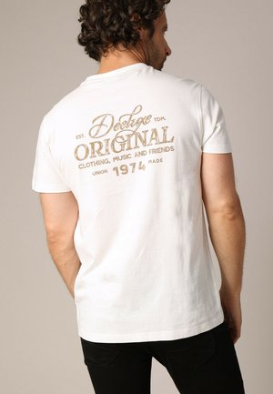 Witte katoenen t-shirt met een decoratieve print op de achterkant in bruin, met de tekst "Deluxe Original" en details over de kleding en oprichtingsjaar.