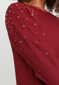 Top bourgogne à manches longues avec des embellissements dorés dispersés, en tissu doux et à coupe décontractée. Texture lisse sur l'ensemble.