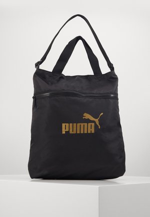 Czarna torba typu tote Puma ze złotym logo, dwoma uchwytami i poziomą kieszenią na zamek, prezentowana na białym piedestale na jednolitym tle.