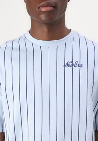 Svetlo modra majica s vertikalnimi temno modrimi črtami, okroglim izrezom, kratkimi rokavi in izvezenim logotipom "New Era" v temni modri barvi na prsih.